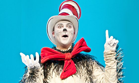 The Cat in the Hat, with his trademark red bow and red and white striped hat, points a finger to the sky like he has an idea.