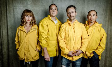 All four band members, one woman and three men, stand against a green curtain. They all wear yellow raincoats and jeans.