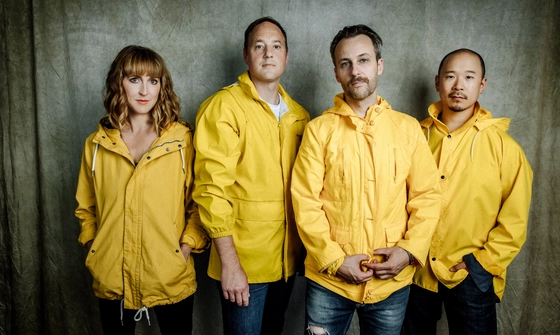 All four band members, one woman and three men, stand against a green curtain. They all wear yellow raincoats and jeans.