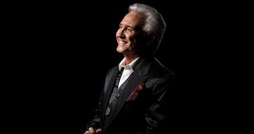 Tony Christie and older man with grey hair sits smiling. He's wearing a black suit and white shirt and is against a black background.