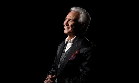 Tony Christie and older man with grey hair sits smiling. He's wearing a black suit and white shirt and is against a black background.