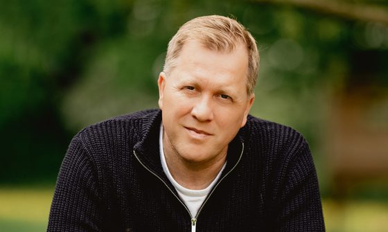 Tony Stockwell is pictured outside in the countryside, wearing a black quarter zip jumper and a white t-shirt
