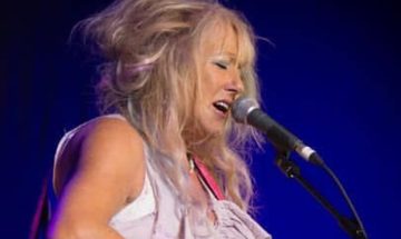 Vikki Clayton, sings into a microphone that is held in a stand. She has long blonde hair and is wearing a white tank top
