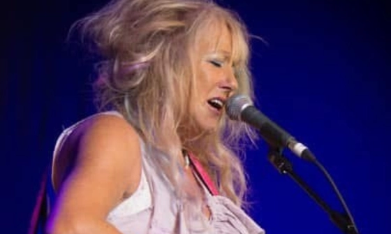 Vikki Clayton, sings into a microphone that is held in a stand. She has long blonde hair and is wearing a white tank top