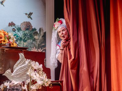 Wendi Peters hiding in a large red curtain and sticking her head out pulling a face. She is wearing the same outfit and headdress from before but now we can see it has large flowers in the front