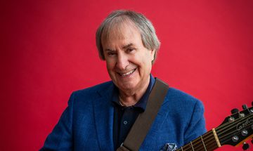 Chris de Burgh, an older white man with long grey/white hair smiling at the camera holding a guitar. He is wearing a black shirt and blue blazer on a red background