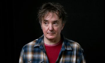Dylan Moran, a white man with scruffy brown greying hair smirking slightly and looking to the left in a red t-shirt with a plaid blue, white, and brown shirt over the top