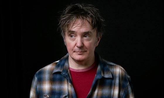 Dylan Moran, a white man with scruffy brown greying hair smirking slightly and looking to the left in a red t-shirt with a plaid blue, white, and brown shirt over the top