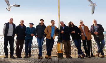 Nine men in casual dress, all with coats, stood be the sea front on what looks to be a pier, two in the centre are leaning on a lamp post with two seagulls flying above them.