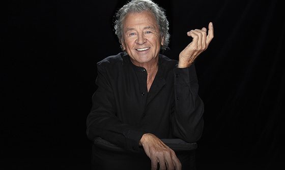 Ian Gillan wearing a black shirt and smiling in front of a black background.