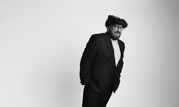 Black-and-white portrait of comedian John Kearns wearing a suit and his signature messy black wig, leaning forward to one side against a plain background.