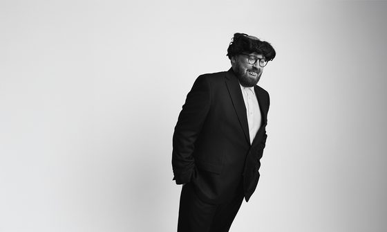 Black-and-white portrait of comedian John Kearns wearing a suit and his signature messy black wig, leaning forward to one side against a plain background.