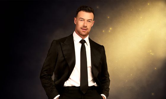 Kai Widdrington, a white man with short dark hair and a short stubbly beard wearing a black suit and tie with a white shirt on a dark background with a golden shaft of light