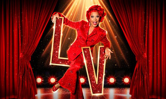 A photo of La Voix, drag queen in a bright red wavy wig and a red quilted suit with diamonte lips covering it. She is leaning on sparkly gold and red letters LV on a stage with red velvet curtains