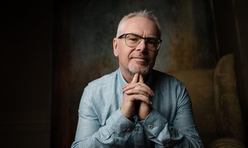 Nik Kershaw sat with his hands clasped together and index fingers under his chin, smirking at the camera.