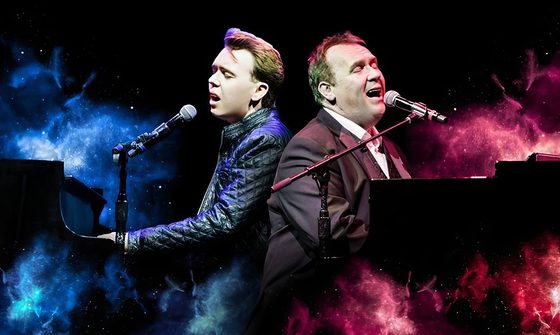 Two white men, sat back to back at pianos playing and singing into microphones with colourful smoke around them. On the left is a young man with dark quiffed hair wearing a leather jacket, he is surrounded by blue smoke. On the right is an older man in a dark suit jacket with red smoke around him