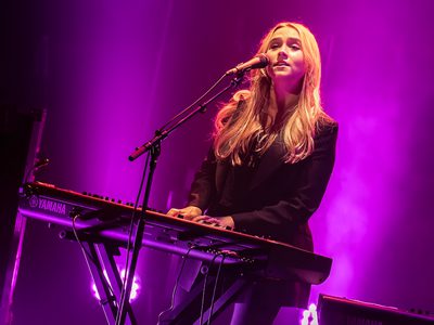 A blonde woman in a black outfit playing keyboard and singing into a microphone under purple lighting.