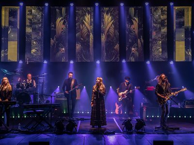 7 band members on stage under spotlights. They are playing guitars, drums, and keyboards, and there are two singers towards the front of the stage, with one playing a keyboard.