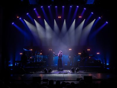 A woman singing on stage under bright spotlights, with a live audience below and a band performing behind her.