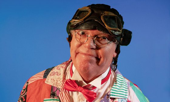 An old man in a pilots hat with goggles on top, round glasses, and a colourful patchwork jacket with a shirt and bow tie smirking at the camera