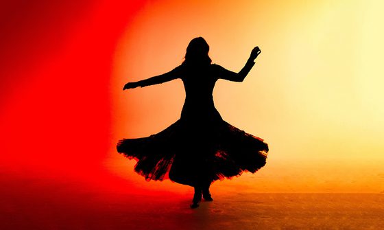 A silhouette of a woman wearing a long, frayed skirt dancing against a luminescent orange background.