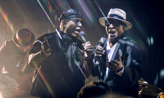 Chris Amoo and Dave Smith of The Real Thing singing on stage together with a man behind playing guitar. The room is dark and the lights are creating a lens flare on the image