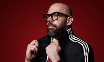 Tom Davis, a bald white man with a dark beard and thick rimmed black glasses. He is holding the flicked up collar of his black jacket with white stripes as he looks at the camera on a red background