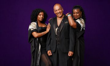 A man in a black suit stands alongside two women in black dresses, all smiling at the camera against a solid purple background.