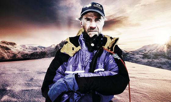 Sir Ranulph Fiennes stood outdoors wearing a black and blue coat with expedition equipment attached, on a dark and cloudy mountainous background