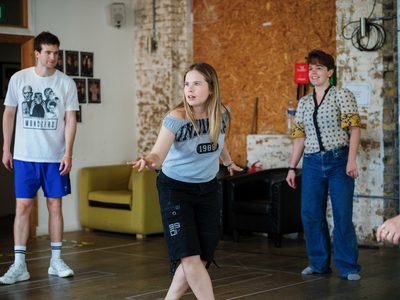 Toby Beynon, Janita Mikkola and Emma Wilkinson rehearse a scene, with Janita in the centre in a crouched pose while the others face her in a circle.