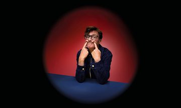 Ed Byrne, a white man with short quiffed brown hair and glasses, poking both of his index fingers into the corners of his mouth making an odd smile