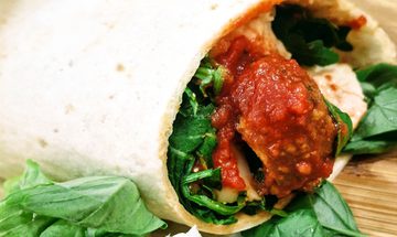 A meatballs in a wrap on a wooden board. The wrap has meatballs, red sauce, basil, and mozzarella