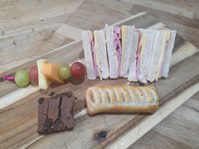 a platter of ham and cheese sandwiches, a sausage roll, fruit and chocolate brownie